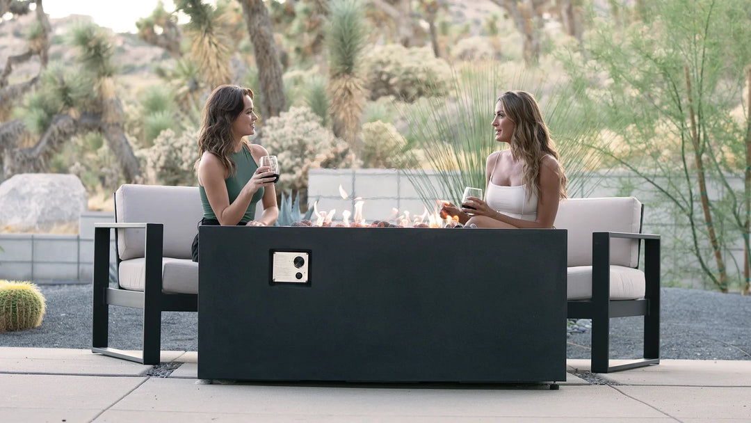 Outdoor patio featuring a sleek black rectangle concrete fire pit with hidden propane tank, set amidst a natural desert landscape. Two women sit on modern outdoor lounge chairs, enjoying drinks and conversation around the warm flames of the fire pit.