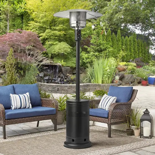 A single standing silver and black patio heater in front of a simple yet elegant patio furniture set with a woodland background.