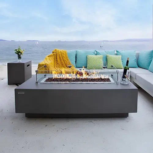 A concrete gas fire pit is resting on a beach front patio surrounded by a comfortable sectional couch with blue pillows.