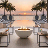 At a waterfront pool deck during sunset, a low white fire bowl acts as a round fire pit between modern lounge chairs and small metal side tables holding drinks, towels, and candles, its flames flickering over lava rocks while palm trees in large planters frame an infinity pool that appears to merge with the calm water beyond, illustrating an outdoor fire pit setting arranged for relaxed resort-style outdoor entertaining.