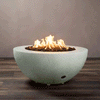 A round, half-sphere fire bowl made of light gray textured concrete rests on a dark wood-look floor, its wide rim framing a recessed basin filled with chunky brown lava rocks while bright flames from the gas fire pit rise and curl in the center, clearly showing the smooth curve of the concrete fire pit, the low metal feet lifting it slightly off the ground, and a small control opening near the base against a plain taupe wall.