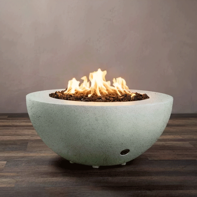 A round, half-sphere fire bowl made of light gray textured concrete rests on a dark wood-look floor, its wide rim framing a recessed basin filled with chunky brown lava rocks while bright flames from the gas fire pit rise and curl in the center, clearly showing the smooth curve of the concrete fire pit, the low metal feet lifting it slightly off the ground, and a small control opening near the base against a plain taupe wall.