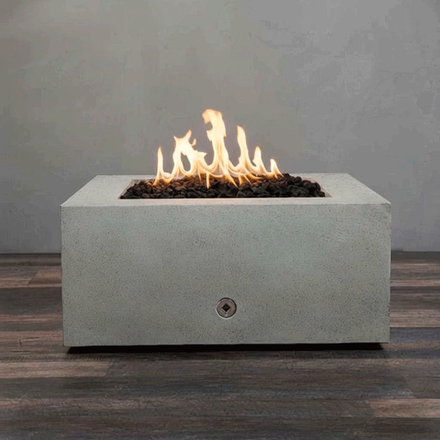 A low, rectangular concrete fire pit with a smooth pale gray surface fills the frame, its recessed center packed with dark lava rocks as a line of tall yellow flames flickers and shifts, the square fire pit resting on wide wood-look planks against a plain gray wall, highlighting the clean, modern shape of this concrete fire pit and the quiet glow of a compact gas fire pit styled as a simple fire table.