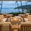 On a covered balcony overlooking a calm ocean and swaying palm trees, a white concrete fire pit flickers at the center of cream cushioned sofas and loungers, its flames lifting from dark stones while side tables hold drinks and orchids, creating a low rectangular fire table that reads as a luxury fire pit in a serene outdoor lounge arranged for relaxed evening entertaining by the water.
