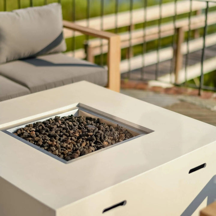 A close-up view of the 42" Square Beton Concrete GFRC Gas Fire Pit by Starfire Designs in a sleek white finish, shown without the flame to highlight its modern design and natural black lava rock media. The smooth GFRC surface and integrated propane tank storage create a clean, contemporary look, perfect for coastal decks, patios, or backyard lounging areas. This image showcases the fire pit's premium craftsmanship and durable materials, ideal for homeowners seeking a minimalist outdoor heating solution.