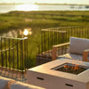 A crisp white 42-inch square Beton concrete gas fire pit by Starfire Designs, featuring a hidden propane tank and modern lava rock burner, radiating a cozy flame as it anchors a stylish outdoor seating area. The serene waterfront view and golden hour glow highlight the luxurious yet inviting atmosphere / perfect for evening relaxation or entertaining on the patio.