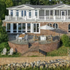 Aerial view of a stunning brick patio overlooking the water, featuring the 42" Square Beton White Concrete GFRC Gas Fire Pit by Starfire Designs. The fire pit is beautifully centered beneath a large outdoor umbrella, surrounded by modern teak and cushion lounge seating, creating a serene and stylish outdoor living space perfect for entertaining or relaxing by the shore.