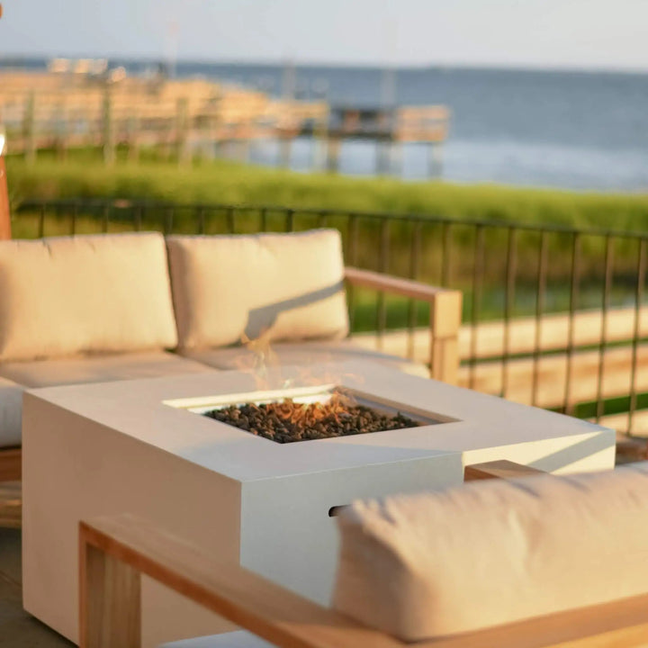 Modern outdoor patio featuring a 42-inch square white GFRC concrete gas fire pit by Starfire Designs, with lava rock media and a hidden propane tank, set against a peaceful waterfront backdrop with warm, cushioned teak lounge seating. Perfect for creating a cozy and stylish backyard retreat.