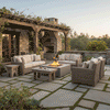 A rustic Tuscan-style courtyard features a brown backyard fire pit surrounded by stone pavers and wicker seating. An outdoor fireplace and lush greenery frame the scene, creating a cozy family gathering fire pit area. The warm glow of the fire feature illuminates the textured stone patio, serving as an inviting outdoor fire pit centerpiece for the garden estate.