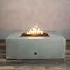 A studio close-up features a square fire pit in a smooth natural gray finish, resting on a dark wood floor. Bright yellow flames burn centrally from a bed of black lava rocks, highlighting the clean geometric lines of the concrete fire pit. The visible chrome key valve on the side indicates the manual control of this natural gas fire pit unit.