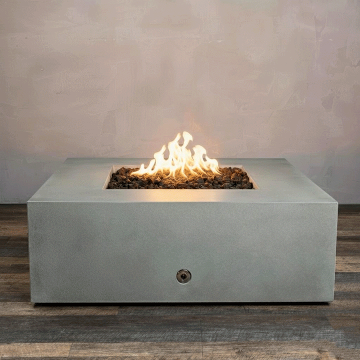 A studio close-up features a square fire pit in a smooth natural gray finish, resting on a dark wood floor. Bright yellow flames burn centrally from a bed of black lava rocks, highlighting the clean geometric lines of the concrete fire pit. The visible chrome key valve on the side indicates the manual control of this natural gas fire pit unit.
