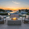 A gray patio fire pit serves as the centerpiece of a waterfront terrace at sunset, surrounded by docked yachts and palm trees. A small sofa and two armchairs frame the square fire pit, creating an intimate setting for outdoor entertaining. The warm glow of the propane fire pit enhances the tranquil atmosphere, establishing a cozy backyard fire pit spot by the water.