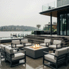 A spacious lakeside terrace features a gray outdoor fire pit surrounded by eight modern armchairs. The scene overlooks a lake and forest on a misty day, highlighting the durability of the concrete fire pit in outdoor elements. The bright flames of the gas fire pit offer a warm contrast to the cool, rainy atmosphere, demonstrating the utility of a smokeless fire pit for year-round enjoyment.
