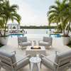 A white luxury fire pit sits centrally on a stone terrace, framed by four armchairs and an infinity pool overlooking a tropical waterway. The steady flames of the modern fire pit reflect on the water's surface, creating a sophisticated atmosphere for outdoor entertaining. The scene highlights the sleek design of this backyard oasis against a serene waterfront backdrop.