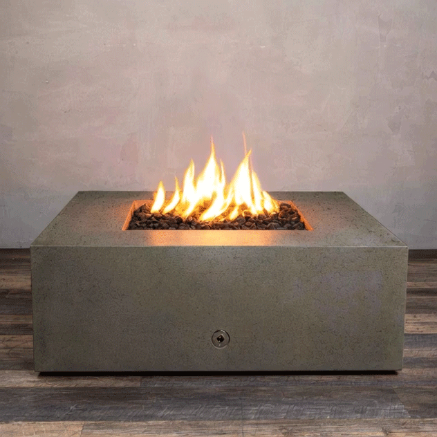 A low brown concrete fire pit with crisp edges sits centered on a striped wood-look floor in front of a soft taupe wall, its recessed square opening piled with uneven dark lava rocks while tall yellow flames from the gas fire pit rise and bend at the center, clearly outlining the broad top surface, front control port, and compact proportions of this modern square fire pit as it burns steadily in place.