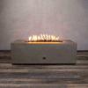 A studio view presents a rectangular concrete fire pit in a natural brown finish, resting on a dark wood floor against a textured beige wall. Bright orange flames rise continuously from a bed of black lava rocks along the length of the linear fire pit. The minimalist backdrop emphasizes the earth-tone texture and clean operation of a natural gas fire pit connection.