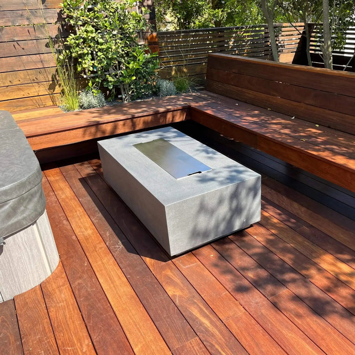 Modern rectangular GFRC gas fire pit in natural gray by Starfire Designs, placed on a rich wood deck patio with wraparound wood bench seating, shown with stainless steel burner cover in place / perfect for a clean and stylish backyard setup.