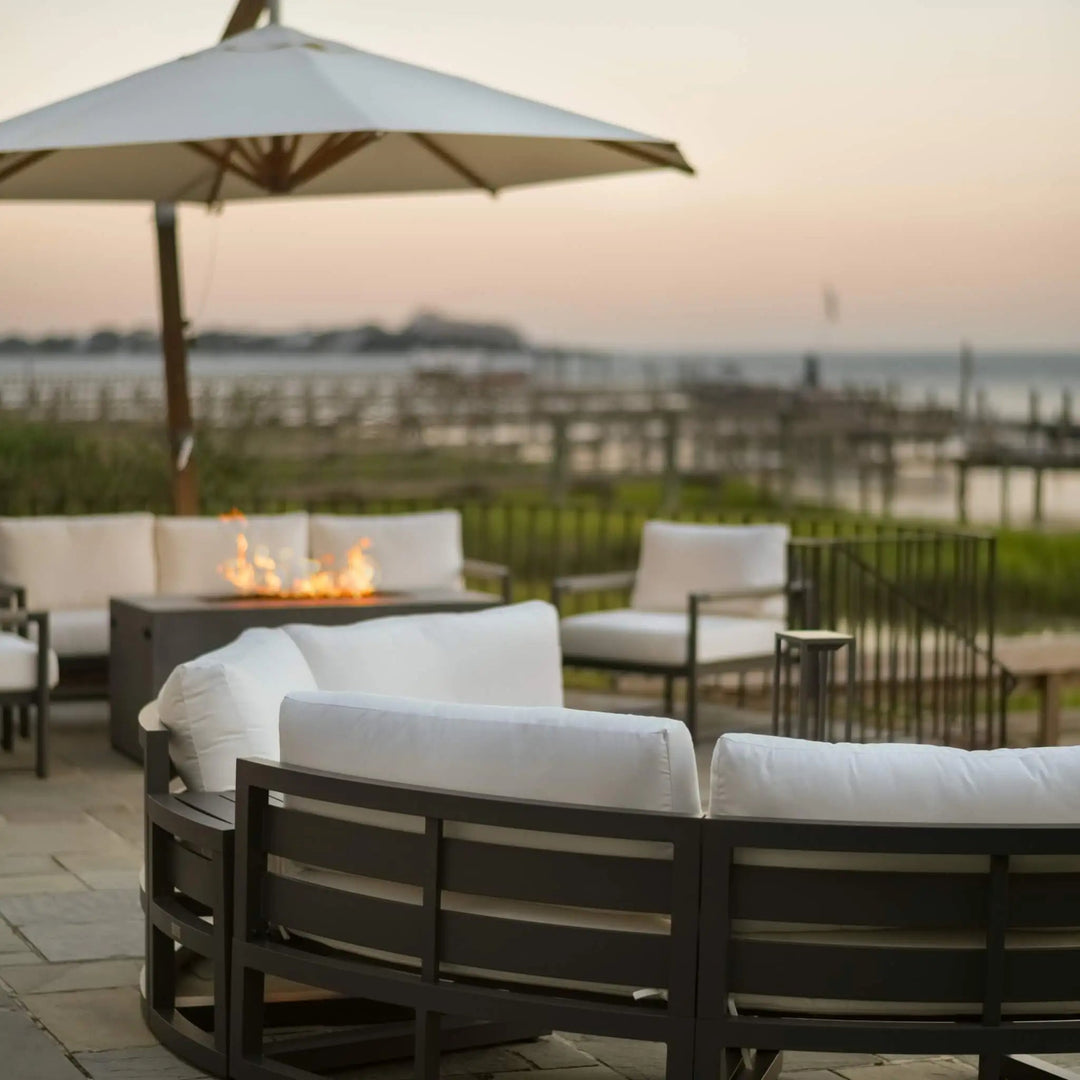 Modern outdoor patio scene featuring a 55-inch rectangular Beton concrete GFRC gas fire pit by Starfire Designs in a sleek graphite finish. The fire pit's clean, contemporary design pairs seamlessly with plush white-cushioned outdoor seating arranged in a semi-circle, all set under a large umbrella with a calming coastal view and wooden dock in the background. Flames gently flicker from the fire pit, creating an inviting ambiance perfect for evening relaxation or waterfront entertaining.