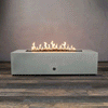 A studio presentation displays a rectangular fire pit in a natural gray finish, resting on a dark wood floor against a grey textured wall. Bright yellow flames burn steadily along the linear fire pit burner, rising from a bed of black lava rocks. The minimalist design highlights the smooth texture of the concrete fire pit, showcasing the clean operation of a natural gas fire pit connection.