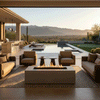 In a desert-style outdoor living room overlooking a pool and distant mountains at golden hour, four cushioned armchairs and two small accent tables gather on a patterned rug around a sand-colored concrete fire pit, its elongated rectangular fire channel glowing with even flames, while cacti, ornamental grasses, and a long bar with stools frame the scene, turning the space into a relaxed backyard fire pit setting that feels like a refined backyard oasis.