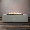 A studio presentation highlights a substantial rectangular fire pit in a natural gray finish, resting on a dark wood floor. Bright yellow flames dance continuously along the linear fire pit burner, rising from a bed of black lava rocks. The minimalist setting emphasizes the robust profile and clean operation of this concrete fire pit, designed for a natural gas fire pit connection.