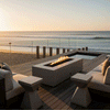 A modern fire pit with a rectangular gray profile sits on a wooden deck next to a small pool, overlooking the ocean at sunset. The setting features a glass railing and sandy beach, creating a tranquil backyard oasis. The bright, steady flames of the fire feature enhance the coastal atmosphere, utilizing a gas fire pit for effortless enjoyment.