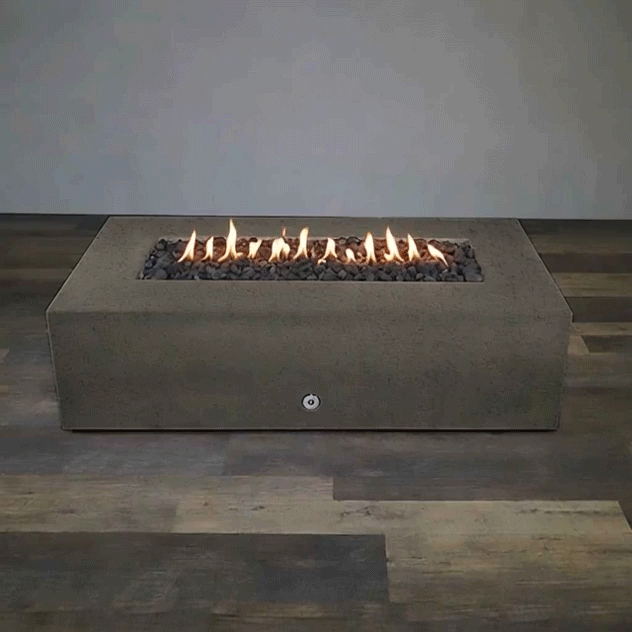 A low brown concrete fire pit with clean rectangular lines sits centered on wood-look flooring in a bare studio, its recessed channel packed with uneven dark lava rocks while a continuous row of yellow flames ripples along the burner, emphasizing the simple boxy silhouette of this concrete fire pit and the stretched proportions of a sleek rectangular fire pit designed as a minimalist linear fire pit focal point.