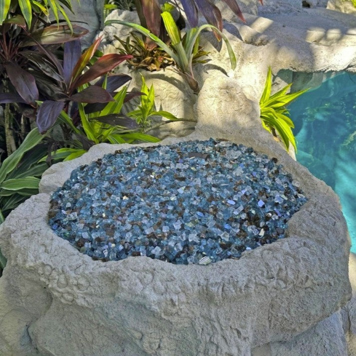 Tropical fire bowl filled with American Fire Products 1/2" Bali pre-mixed reflective fire glass, showcasing a mix of ocean blues and rich bronze tones nestled in natural rock near a poolside oasis.