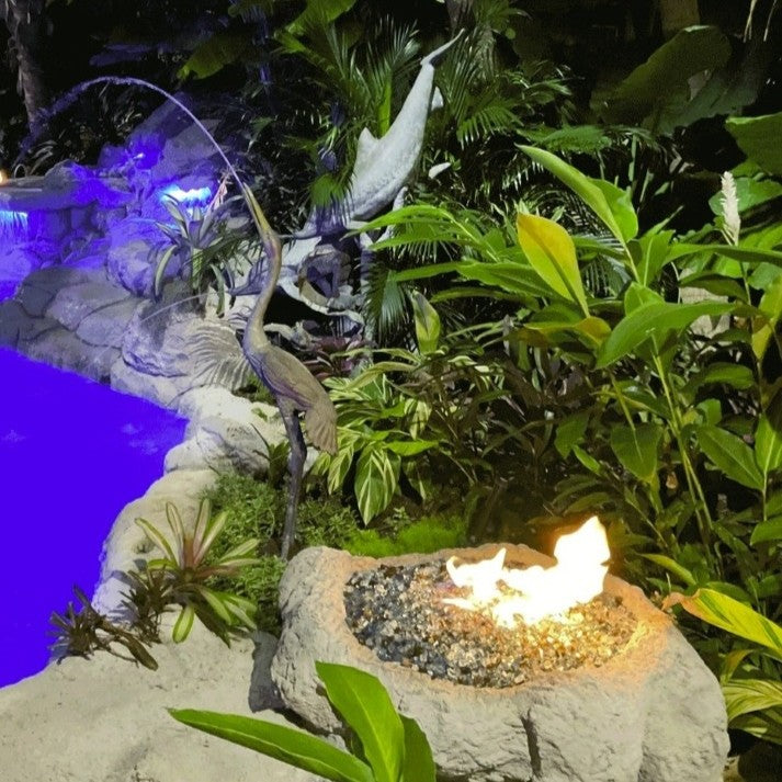 Nighttime tropical fire feature glowing with American Fire Products 1/2" Bali pre-mixed reflective fire glass, radiating warm flames amid lush greenery beside a lit pool and decorative heron statue.
