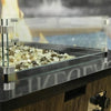 Close-up of a modern fire pit filled with 1/2" Las Vegas Pre-Mix Reflective Fire Glass from American Fire Glass, featuring a stylish blend of gold, bronze, and clear glass chips within a sleek glass wind guard atop a wood-textured base—perfect for luxury outdoor settings.