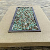 Rectangular fire pit filled with American Fire Glass 1/2" Yosemite Pre-Mixed Reflective Fire Glass, showcasing a natural blend of emerald green, amber, and clear glass pieces—set into a smooth stucco tabletop with a dark metal trim, perfect for outdoor patio gatherings on a paved stone terrace.