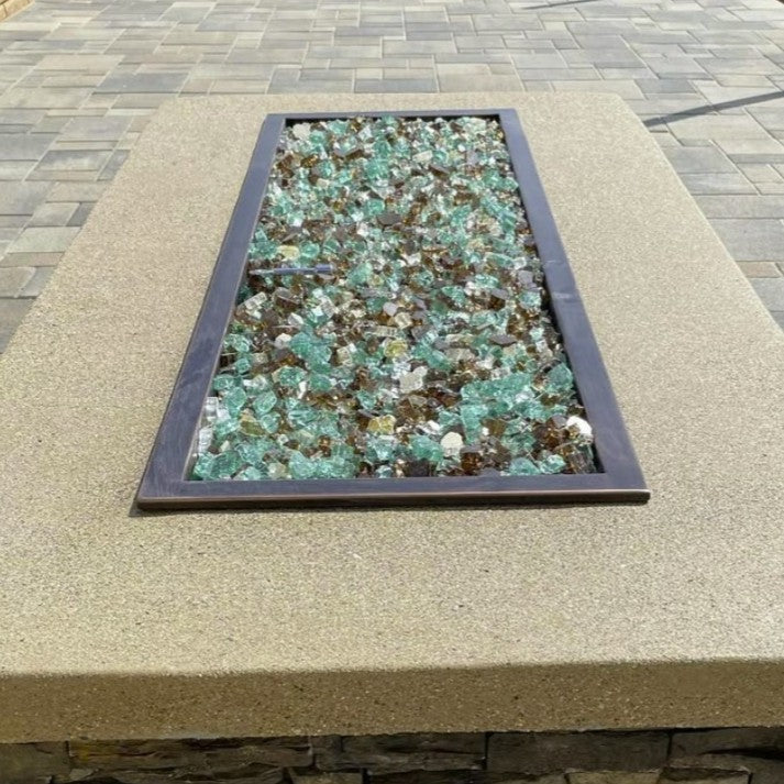 Rectangular fire pit filled with American Fire Glass 1/2" Yosemite Pre-Mixed Reflective Fire Glass, showcasing a natural blend of emerald green, amber, and clear glass pieces—set into a smooth stucco tabletop with a dark metal trim, perfect for outdoor patio gatherings on a paved stone terrace.
