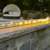 Outdoor fire feature showcasing American Fire Products 1/2" Bora Bora 
reflective pre-mixed fire glass glowing beneath a sleek, linear flame—set atop a stone wall for a luxurious and modern patio ambiance.