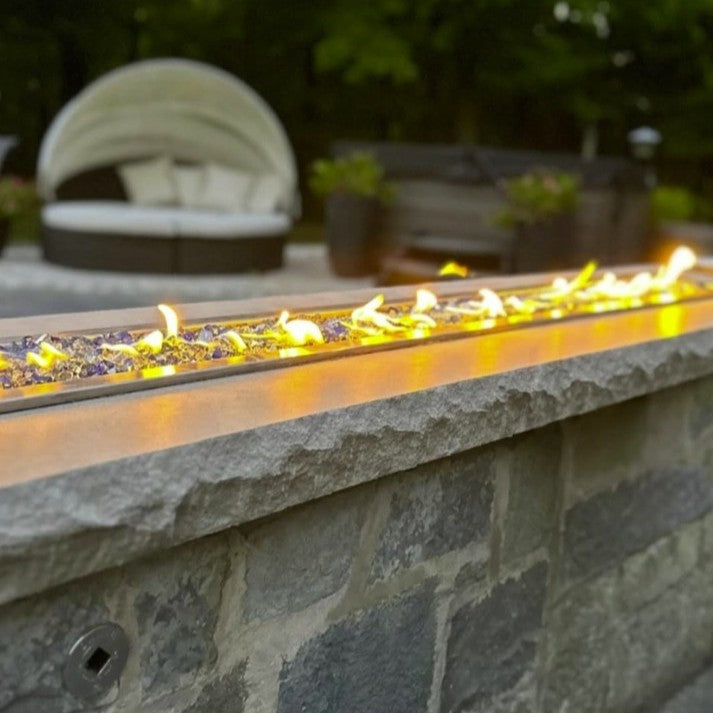 Outdoor fire feature showcasing American Fire Products 1/2" Bora Bora 
reflective pre-mixed fire glass glowing beneath a sleek, linear flame—set atop a stone wall for a luxurious and modern patio ambiance.