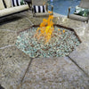 Outdoor octagon fire pit featuring a vibrant flame dancing over a bed of 1/2" Pre-Mixed Reflective Fire Glass from American Fire Glass, with a blend of clear and light aqua tones—set within a polished stone tabletop, surrounded by cozy patio seating near a poolside lounge.