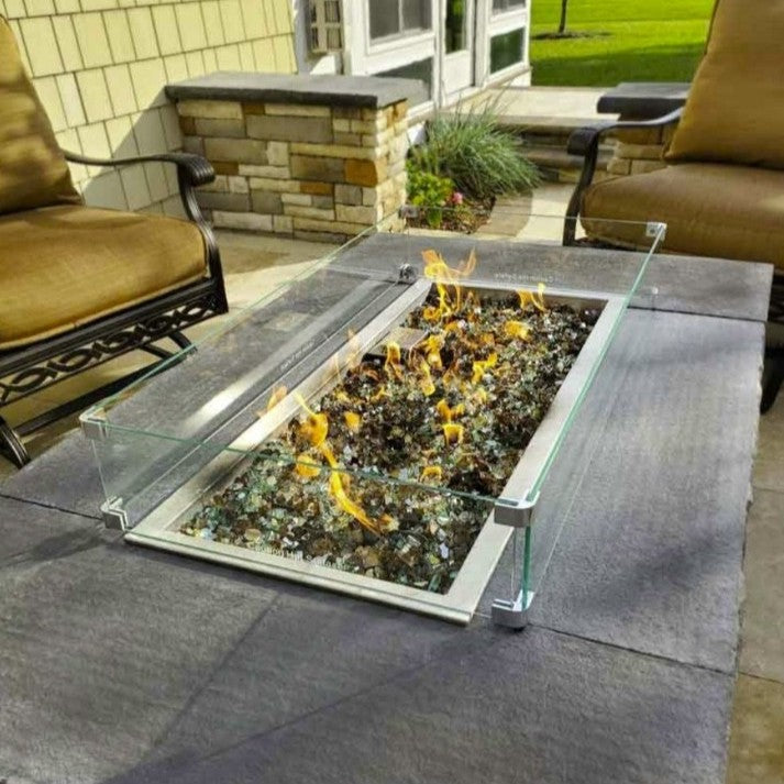 Modern outdoor fire table featuring 1/2" Yosemite Pre-Mixed Reflective Fire Glass from American Fire Glass, with a warm blend of clear, green, amber, and golden glass pieces glowing beneath a lively flame—framed by a sleek glass wind guard and surrounded by comfortable patio seating in a serene backyard setting.