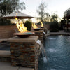 Elegant poolside scene featuring cascading water fountains and raised copper fire bowls filled with American Fire Products 1/4" Non-Reflective Fire Glass, radiating warm flames that create a luxurious and tranquil backyard ambiance at dusk.