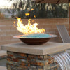 Elegant outdoor fire feature showcasing American Fire Products 1/4" Non-Reflective Classic Fire Glass in a soft aqua hue, beautifully glowing in a copper bowl atop a stone pedestal with integrated water flow—perfect for luxurious backyard ambiance.