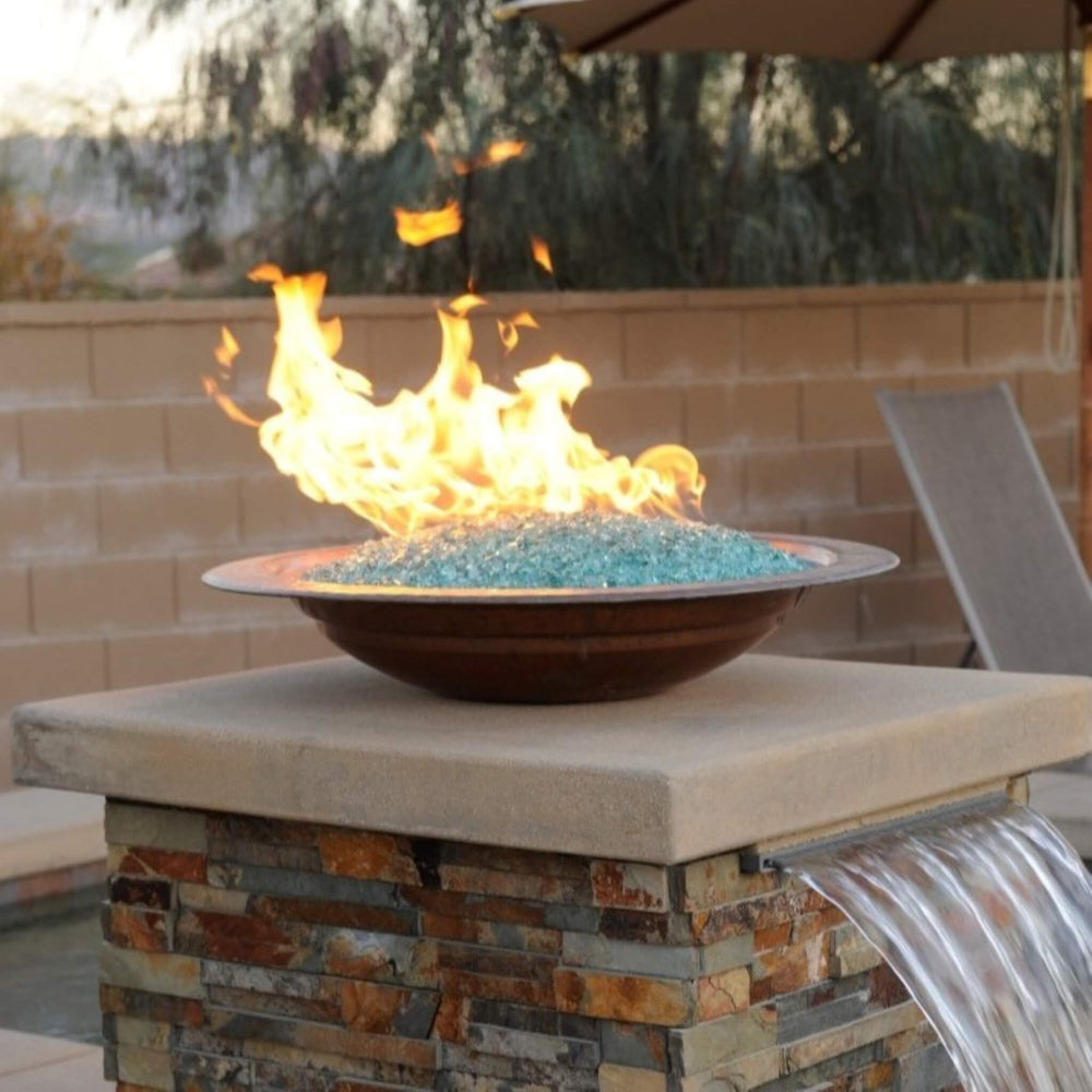 Elegant outdoor fire feature showcasing American Fire Products 1/4" Non-Reflective Classic Fire Glass in a soft aqua hue, beautifully glowing in a copper bowl atop a stone pedestal with integrated water flow—perfect for luxurious backyard ambiance.