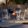 Cozy backyard setup featuring a round stone fire pit filled with reflective Azuria fire glass, producing a tall, elegant flame. Surrounded by classic white rocking chairs and plush seating with striped cushions, this inviting space is framed by a wood fence and tropical greenery—perfect for evening gatherings or peaceful moments by the fire. Available at starfiredirecv.com.