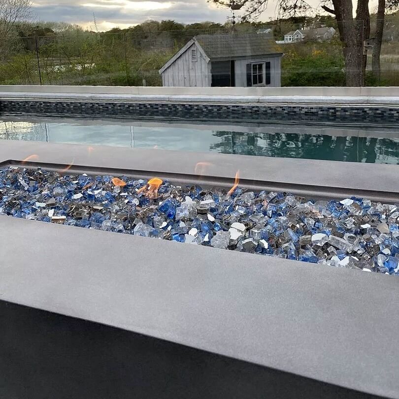 Close-up of a modern rectangular fire pit filled with American Fire Glass 1/2" Maui Breeze pre-mixed fire glass, featuring a sparkling blend of sky blue, cobalt, and silver tones. Set beside a tranquil pool with scenic views, this setup captures the essence of outdoor relaxation and upscale ambiance—available now at starfiredirecv.com.