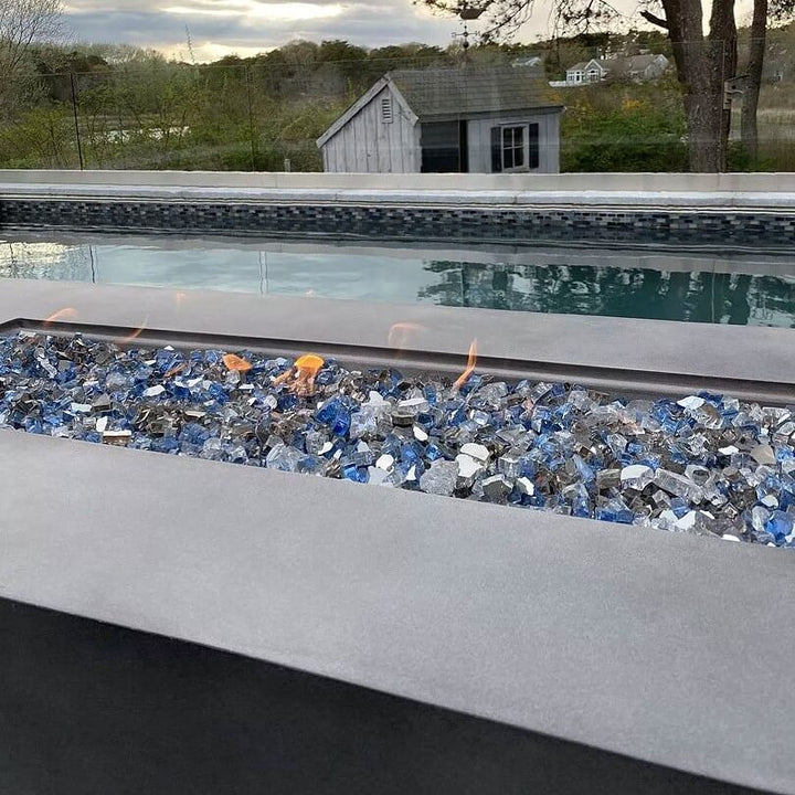 Close-up of a modern rectangular fire pit filled with American Fire Glass 1/2" Maui Breeze pre-mixed fire glass, featuring a sparkling blend of sky blue, cobalt, and silver tones. Set beside a tranquil pool with scenic views, this setup captures the essence of outdoor relaxation and upscale ambiance—available now at starfiredirecv.com.