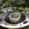 Modern backyard fire pit scene featuring American Fire Products Small Gray Lava Stones in a sleek round bowl, surrounded by curved concrete seating, tropical pillows, lush greenery, and a curious white dog enjoying the cozy outdoor ambiance.