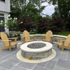 A traditional round stone fire pit filled with American Fire Products Small Gray Lava Stones, surrounded by natural wood Adirondack chairs on a flagstone patio—an inviting backyard gathering space framed by lush greenery and blooming flowers.