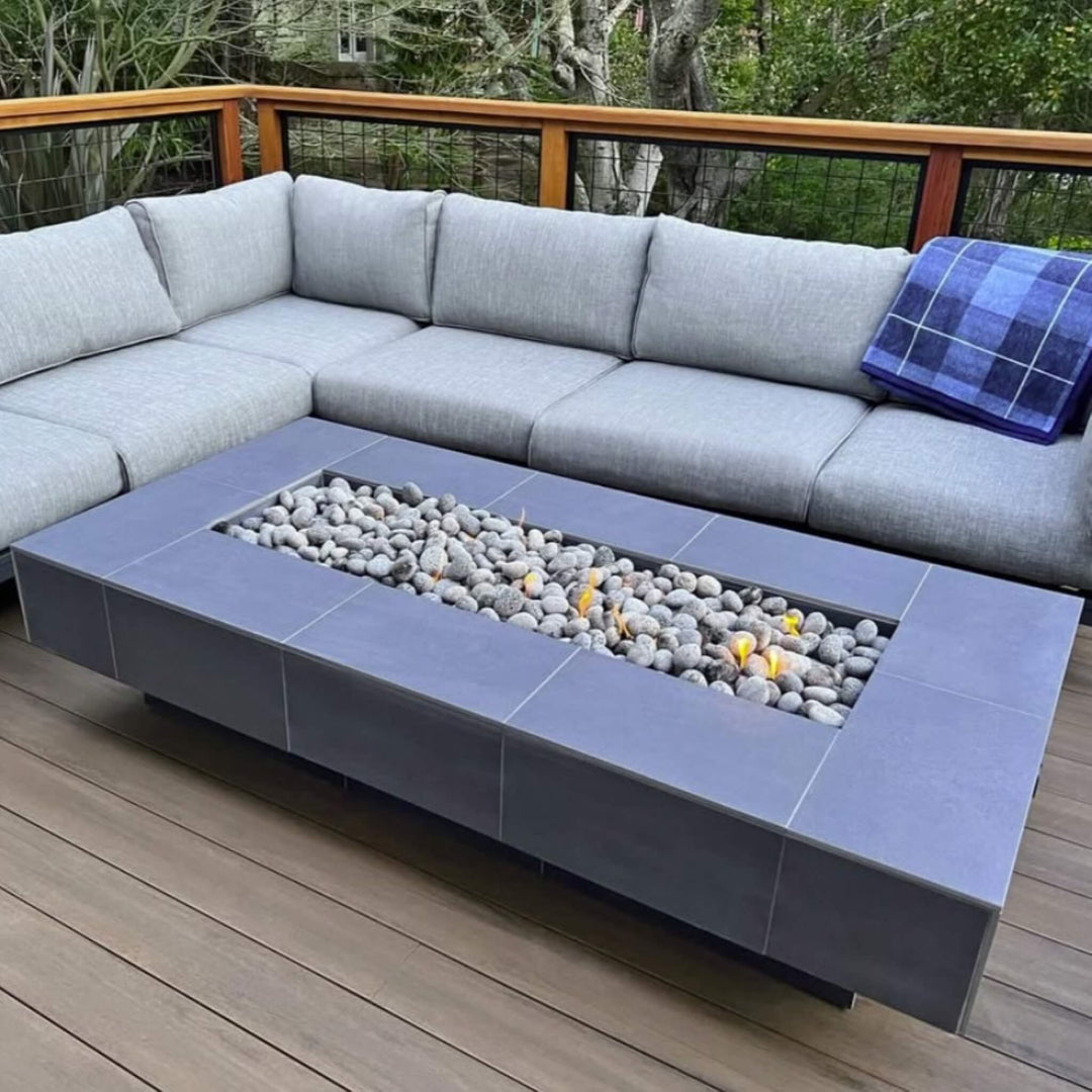 Modern outdoor patio setup featuring a sleek rectangular fire pit table filled with American Fire Products Small Gray Lava Stones, subtly lit by golden flames and paired with a cozy gray sectional for a contemporary, relaxing atmosphere.