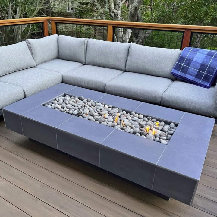 Modern outdoor patio setup featuring a sleek rectangular fire pit table filled with American Fire Products Small Gray Lava Stones, subtly lit by golden flames and paired with a cozy gray sectional for a contemporary, relaxing atmosphere.
