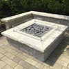Contemporary square fire pit built with stacked stone and topped with American Fire Products Small Gray Lava Stones, enclosed by a clear glass wind guard for added safety and style in a refined outdoor living space.