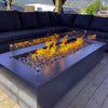 Sleek modern fire table filled with American Fire Products Small Gray Lava Stones, featuring a vivid linear flame behind a clear glass wind guard, surrounded by a deep navy sectional on a wood deck—perfect for upscale outdoor entertaining.