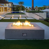 Modern luxury backyard featuring a rectangular concrete fire pit with a brilliant flame powered by an American Fire Products stainless steel drop-in kit. The CSA-certified burner delivers clean-burning fire across a linear tray, surrounded by reflective fire glass for added sparkle. The fire pit sits on a sleek lawn with a pool, palm trees, and contemporary architecture in the background—perfectly blending design, safety, and ambiance.