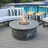 A stylish backyard fire pit setup featuring a round stone fire pit with a smooth modern top, centered around a bright flame fueled by an American Fire Products stainless steel drop-in kit. This CSA-certified system ensures safety while showcasing reflective fire glass. Surrounded by cozy white patio seating and set beside a sparkling pool, the scene blends relaxation, safety, and modern design for the perfect outdoor gathering space.